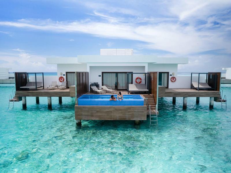 Überwasser-Bungalow mit Terrasse und Sonnenliegen über klarem, türkisfarbenem Wasser.