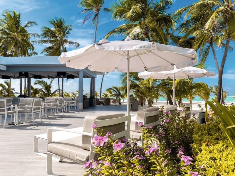 Terrasse mit weißen Sitzmöbeln und Sonnenschirmen, umgeben von Palmen und blühenden Pflanzen.