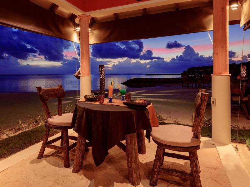 Gemütlicher Essplatz unter Pavillon mit Holzstühlen am Strand bei Sonnenuntergang.