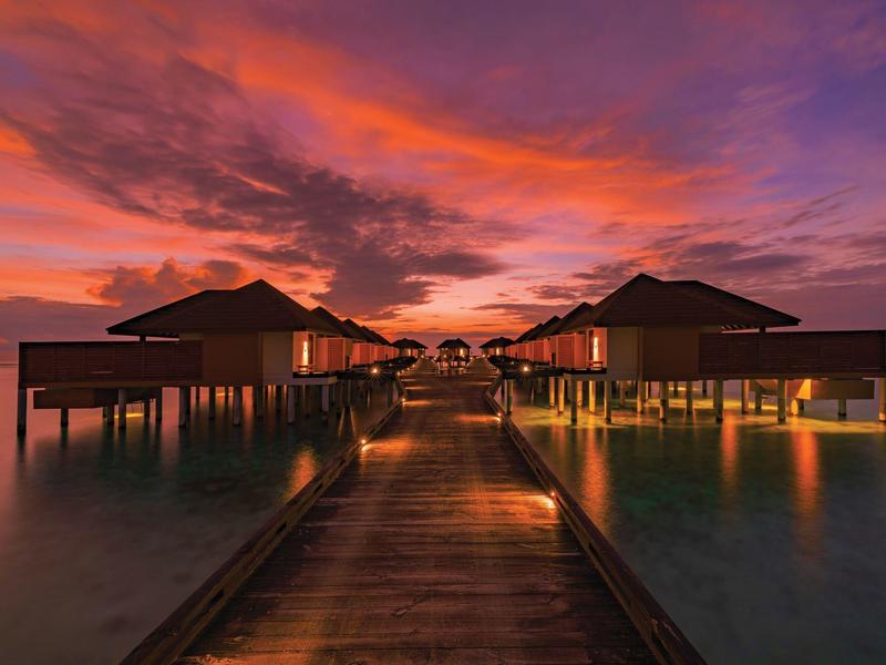 Steg zu Wasserbungalows bei Sonnenuntergang mit orangem und violettem Himmel über ruhigem Meer.