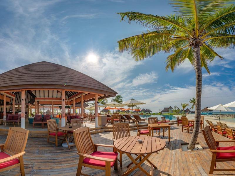 Strandrestaurant mit Holztischen, roten Stühlen, Palmen und Sonnenschirmen bei blauem Himmel.