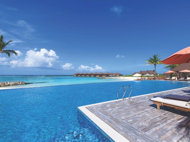 Infinity-Pool mit klarem blauem Wasser, Liegestühlen, Sonnenschirm und tropischem Strand im Hintergrund.