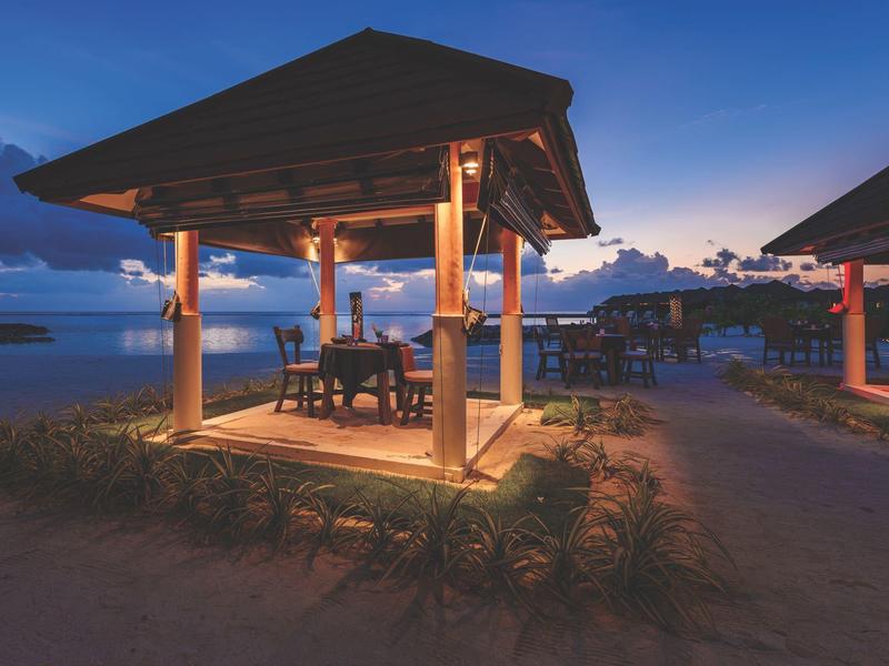 Beleuchtete Holzhütte mit Tisch und Stühlen am Strand bei Dämmerung unter blauem Himmel.