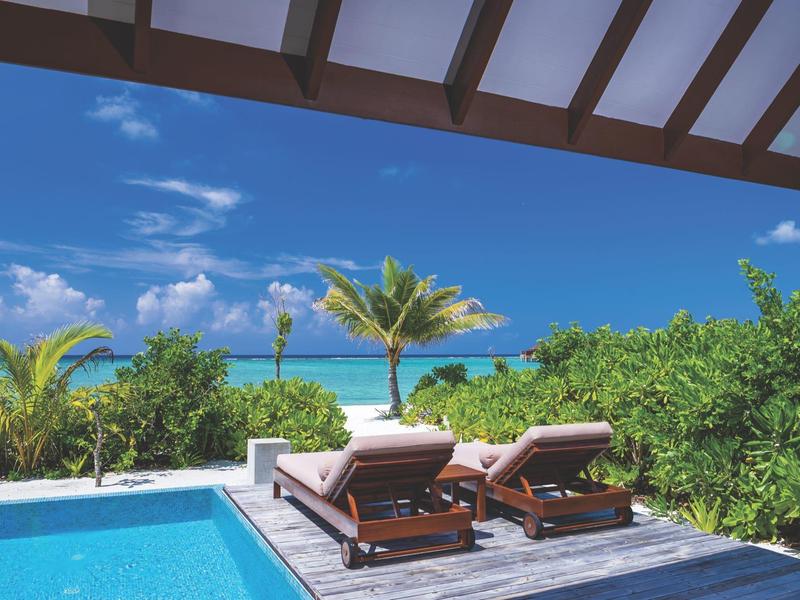 Blick von einer Terrasse mit Pool auf zwei Liegestühle, Palmen und das türkisfarbene Meer unter blauem Himmel.