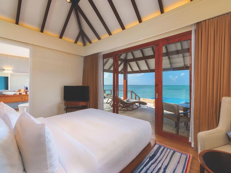 Helles Schlafzimmer mit großem Bett, TV, Sessel und Meerblick durch Glas-Schiebetüren.