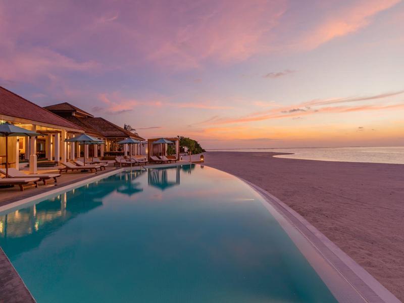Infinity-Pool am Strand mit Häusern, ruhiges Meer und bunter Sonnenuntergang am Horizont.