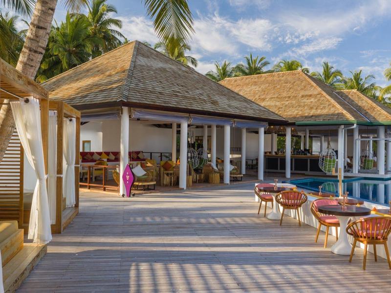 Resort mit Holzboden, Palmendachbungalows, rosa Stühlen und blauem Pool unter blauem Himmel.