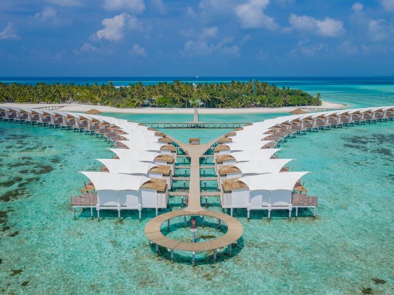 Reihen von Holzhäusern auf Stelzen über klarem, türkisfarbenem Wasser mit Sandinsel im Hintergrund.