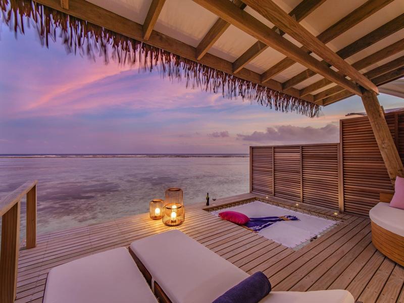 Strandbungalow mit Holzterrasse, Liegestühlen, Kerzen und Blick auf ruhiges Meer bei Sonnenuntergang.