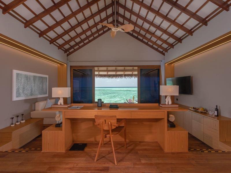 Helles Zimmer mit Holzboden, Schreibtisch, Stuhl, zwei Lampen, großen Fenstern und Deckenventilator.