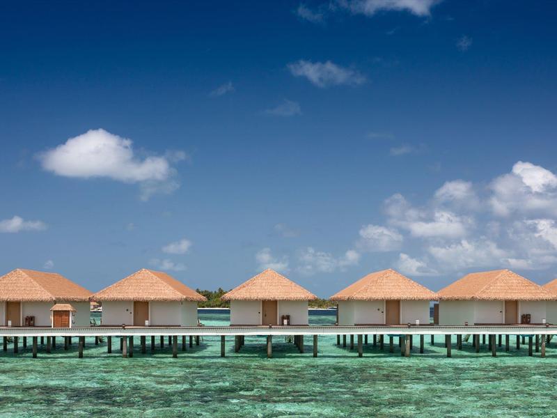 Bungalows auf Stelzen über klarem, türkisfarbenem Wasser unter blauem Himmel mit Wolken.