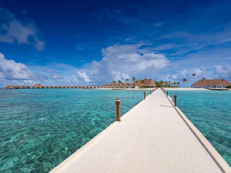 Langer Steg führt über klares türkisblaues Wasser zu kleinen Häusern auf einer tropischen Insel.