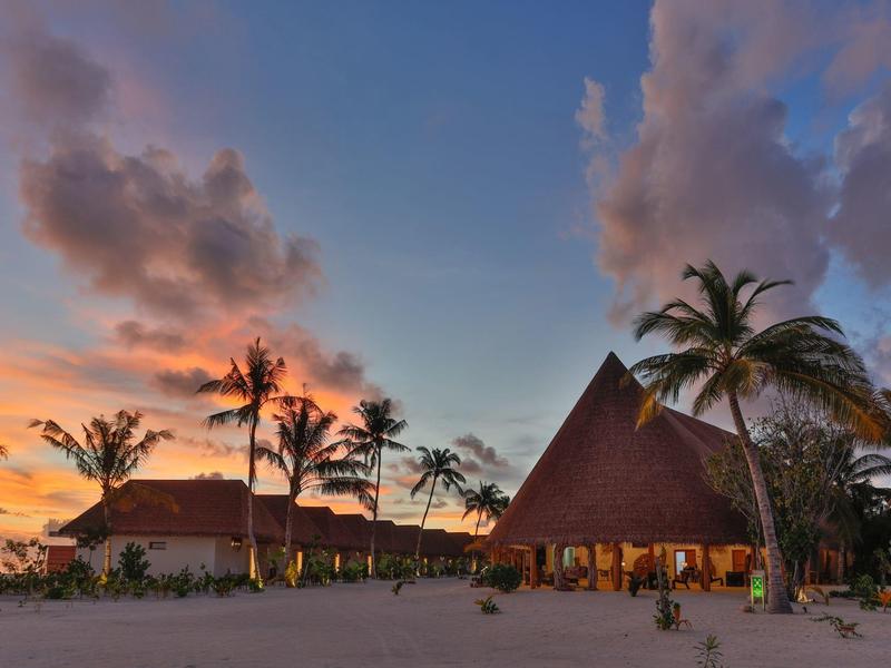 Sonnenuntergang am Strand mit Palmen, Sand und strohgedeckten Hütten unter blau-orangefarbenem Himmel.