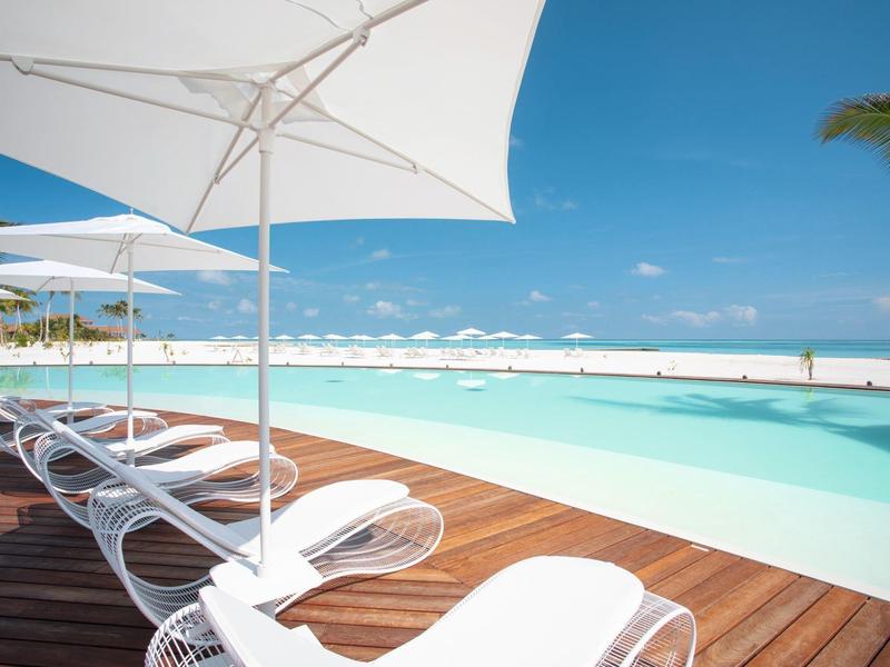 Infinity-Pool mit weißem Sonnenschirm und Liegestühlen auf Holzdeck bei klarem Himmel und Palmen.