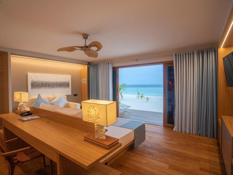 Moderner Hotelzimmer mit Holzmöbeln, Deckenventilator, großer Lampe und Meerblick durch offene Balkontür.