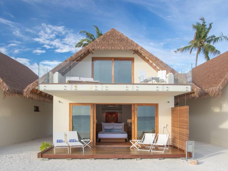 Modernes Strandhaus mit Holzterrasse, zwei Liegestühlen, angrenzendem Sand und blauem Himmel.