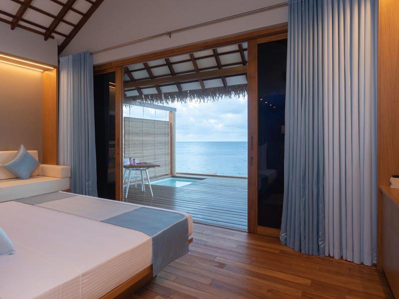 Modernes Hotelzimmer mit Doppelbett, Holzboden und offenem Balkon mit Meerblick und blauem Himmel.