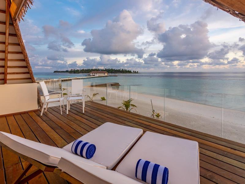 Balkon mit Holzterrasse, zwei weißen Liegen, Stühlen, Blick auf ruhiges Meer und Wolkenhimmel.