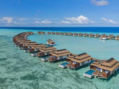 Reihenförmige Wasserbungalows mit Holzterrassen über klarem, türkisfarbenem Meer und blauem Himmel.