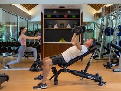 Hombre levanta pesas en un banco inclinado mientras una mujer hace estocadas en un gimnasio.