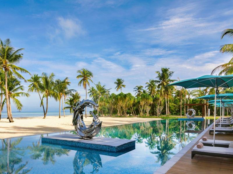 Luxuriöser Pool mit Sonnenschirmen neben Palmen am Strand unter blauem Himmel.