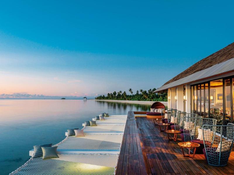 Langer Steg mit Sitzbereich und Pool ins ruhige Meer bei Sonnenuntergang, blauer Himmel, Palmen im Hintergrund.
