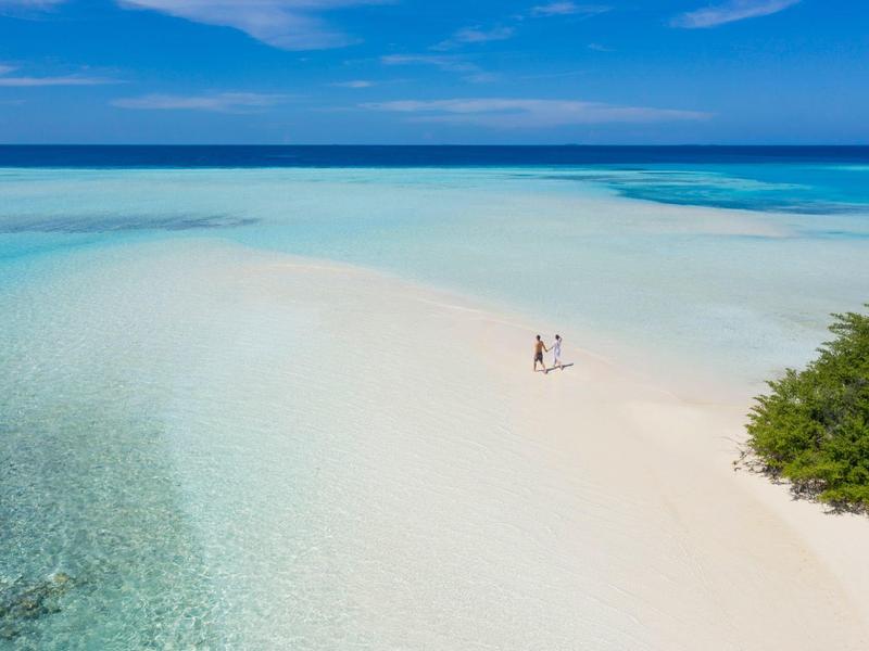 Strand mit weißem Sand, türkisfarbenem Wasser und zwei Personen neben grünem Busch im klaren Himmel.