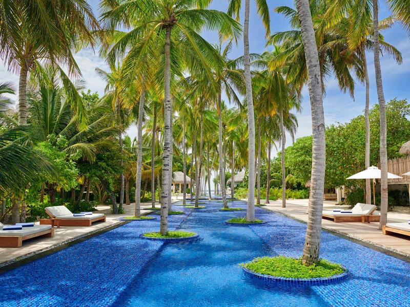 Langer Pool mit blauem Wasser, Palmen auf Inseln und Liegen an den Seiten unter blauem Himmel.