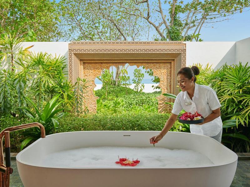 Frau neben großer weißer Badewanne mit Schaum und Blumen, umgeben von grünen Pflanzen und offenem Fenster.