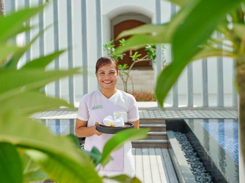 Ein Junge mit braunen Haaren sitzt lächelnd auf einer Holzbank zwischen grünen Pflanzen.