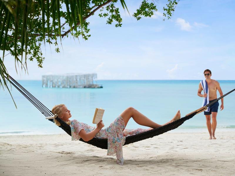 Frau liest entspannt in Hängematte am Strand, Mann lehnt an Stange, Palmenblätter oben.