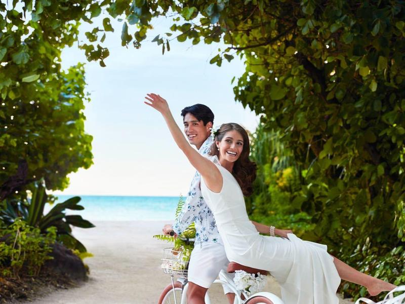 Ein Paar auf weißen Fahrrädern auf einem Weg, umgeben von grünen Bäumen bis zum blauen Meer.