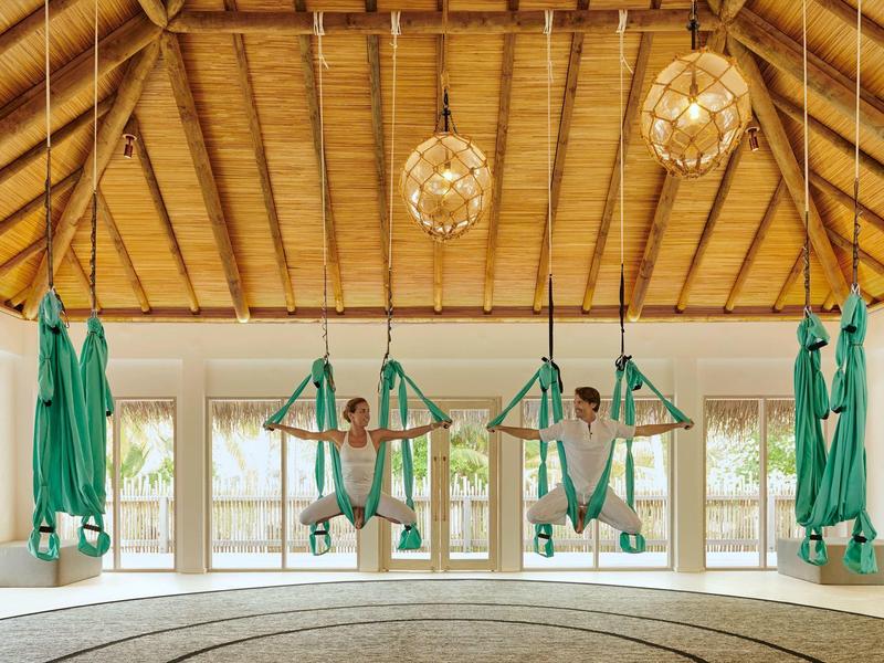 Drei Menschen hängen in grünen Yogatüchern in einer hellen, offenen Halle mit Holzdach und Rundteppich.