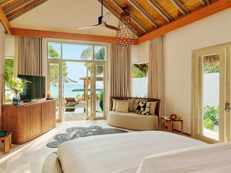 Helles Schlafzimmer mit Holzbalkendecke, großem Bett, Sofa, TV, Balkon und Blick auf Wasser und Natur.