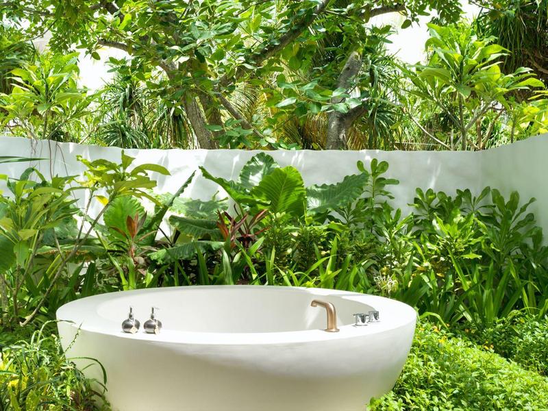 Rundbadewanne im Garten, umgeben von grünen Pflanzen und weißen Wänden bei Tageslicht.