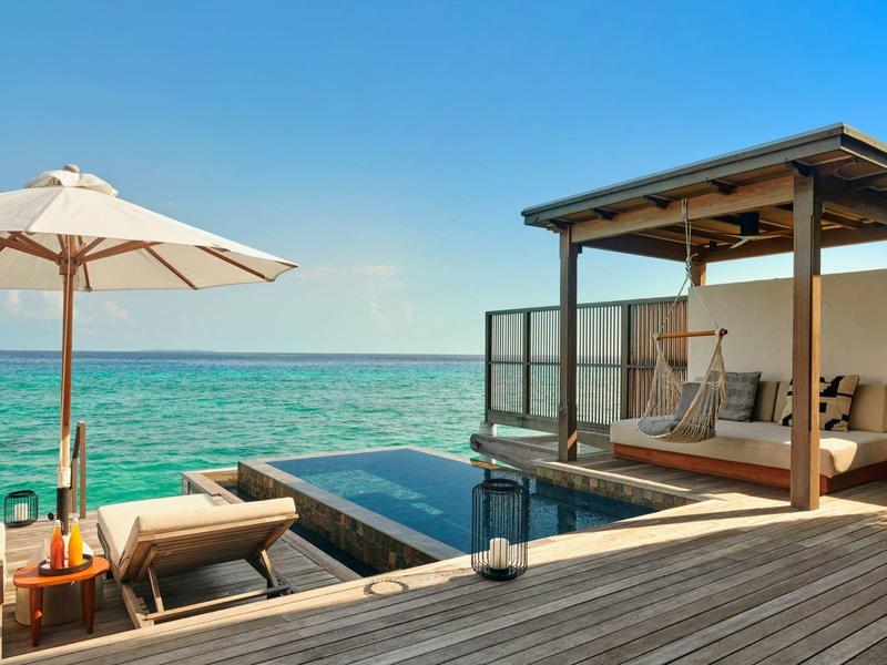 Moderner Holzsteg mit Lounge, Sonnenschirm und Blick auf türkisfarbenes Meer unter klarem Himmel.