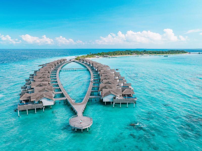 Luxuriöse Wasserbungalows in V-Form über türkisfarbenem Meer unter blauem Himmel.
