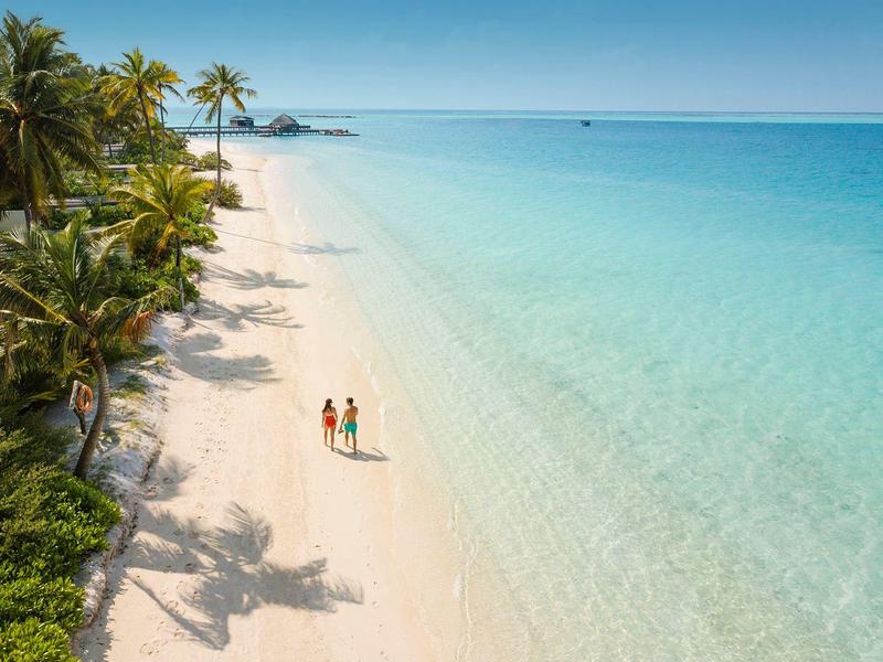 Ein weißer Sandstrand mit Palmen und zwei Personen, die am türkisblauen Wasser spazieren.