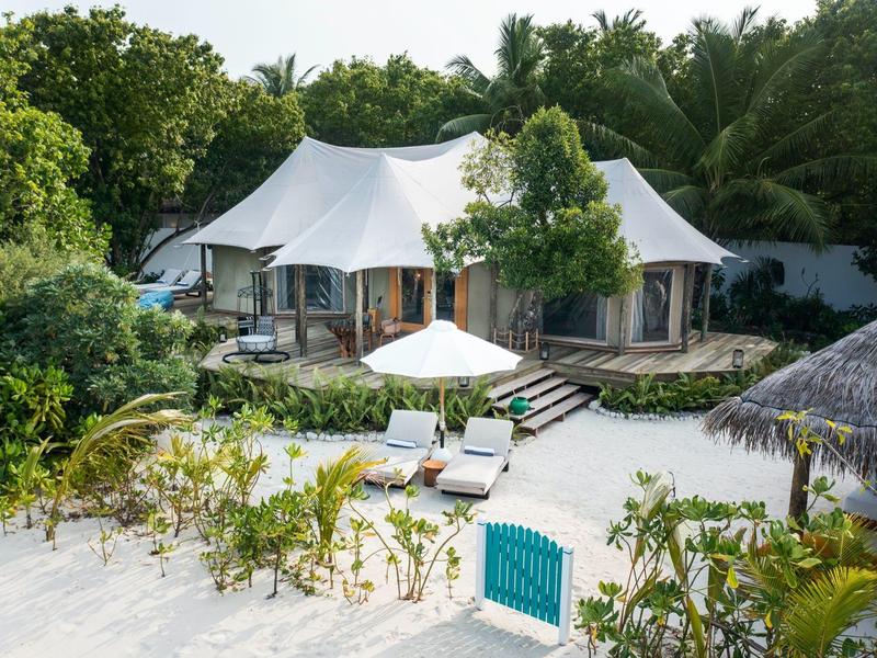 Weiße Strandvilla mit Zeltdach, umgeben von Palmen und grünem Garten, vor weißem Sandstrand.