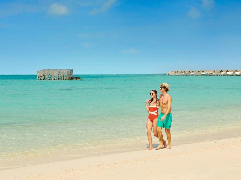Ein Paar in Badebekleidung geht am weißen Sandstrand mit klarem, türkisblauem Wasser und blauem Himmel.
