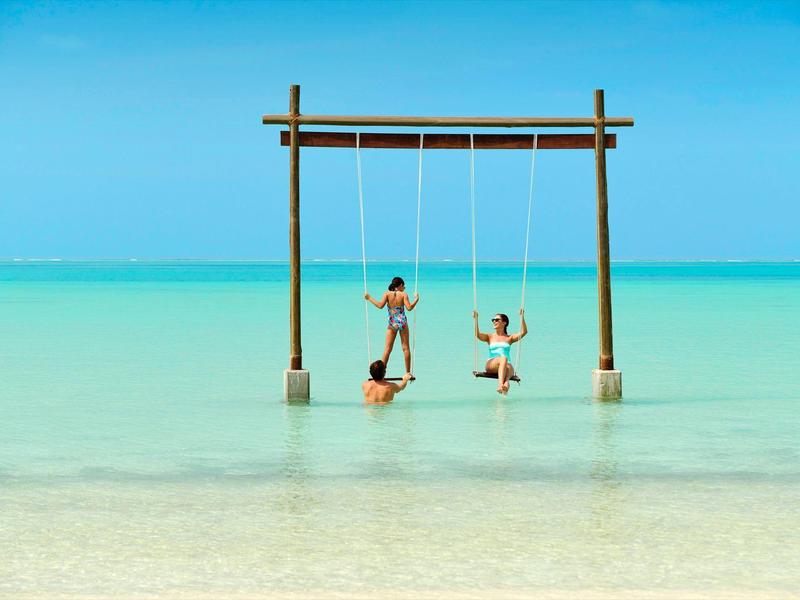 Zwei Kinder schaukeln am Holzgestell im flachen, türkisfarbenen Meer bei blauem Himmel.