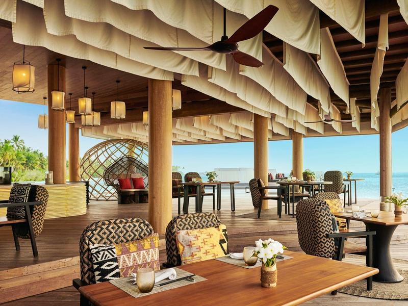 Offener Restaurantbereich mit Holztischen, Rattanstühlen und Blick auf das blaue Meer.