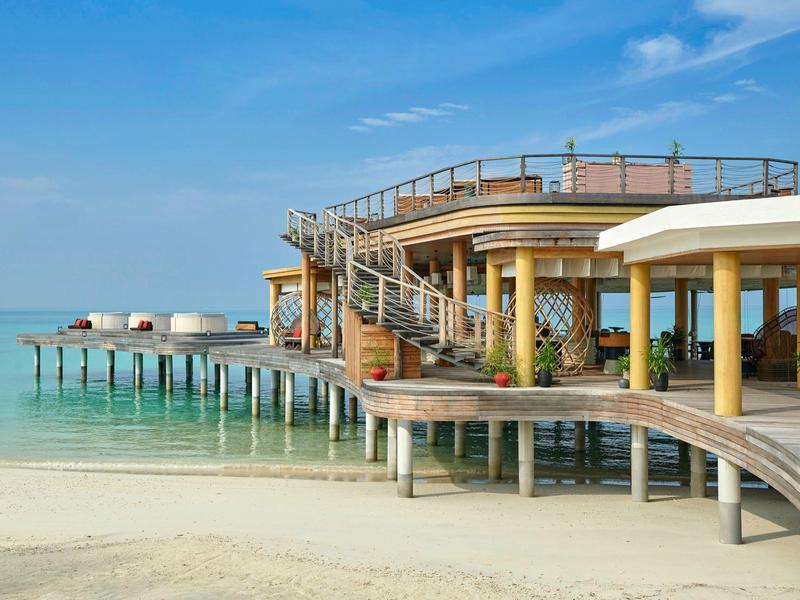 Ein modernes, rundes Strandhaus auf Stelzen mit Steg über klarem, türkisfarbenem Wasser.