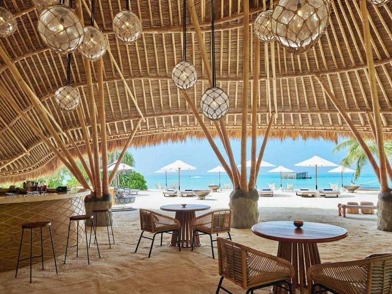 Offene Strandbar mit Rundholzsäulen, Holzdach und weißen Sonnenschirmen am Meer.