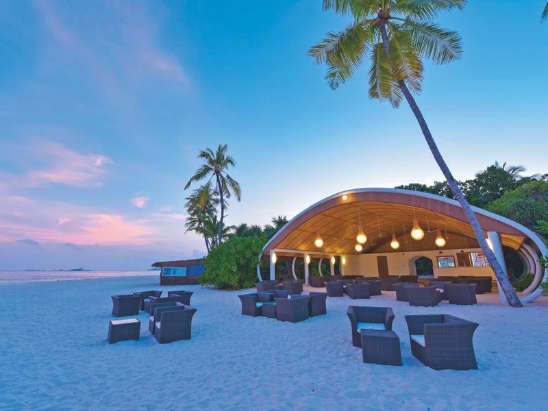 Offener Strandpavillon mit Sitzgelegenheiten und Palmen bei Sonnenuntergang.