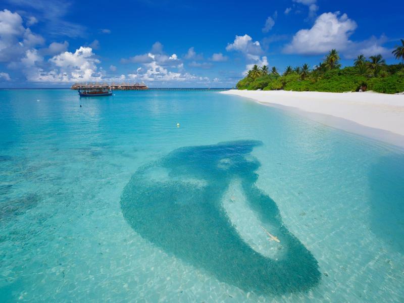 Klares türkisfarbenes Meer mit Sandbank, Schatten eines Mannhechts und blauem Himmel mit Wolken.