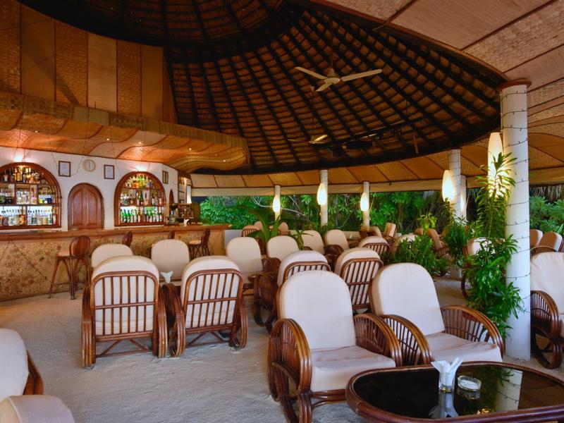 Offener Lounge-Bereich mit Holzmöbeln, weißen Polstern, Pflanzen und geflochtenem Deckendach.