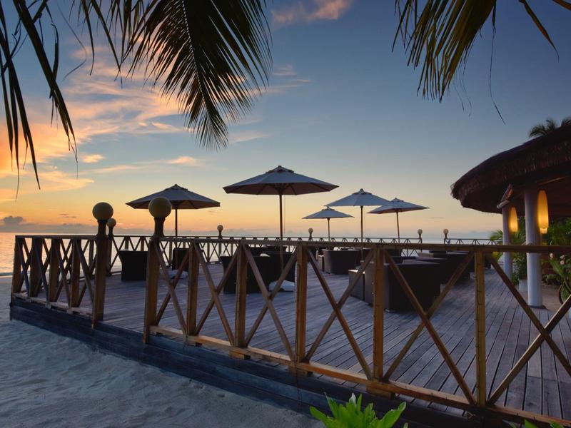 Strand mit Holzgeländer, Sonnenschirmen und Palmen bei Sonnenuntergang am Meer.