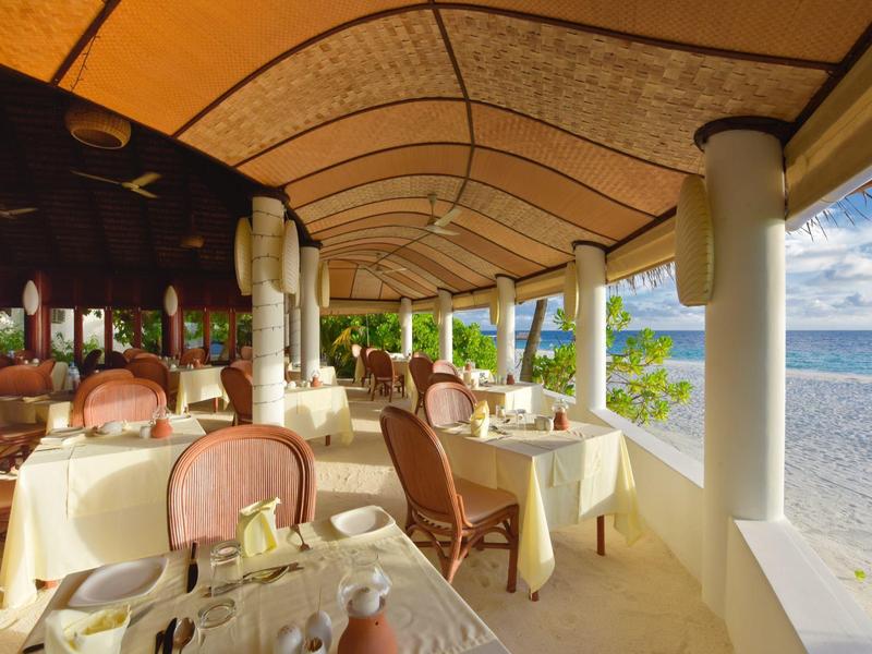 Offenes Restaurant mit gedeckten Tischen, Holzdecke und Blick auf weißen Strand und Meer.