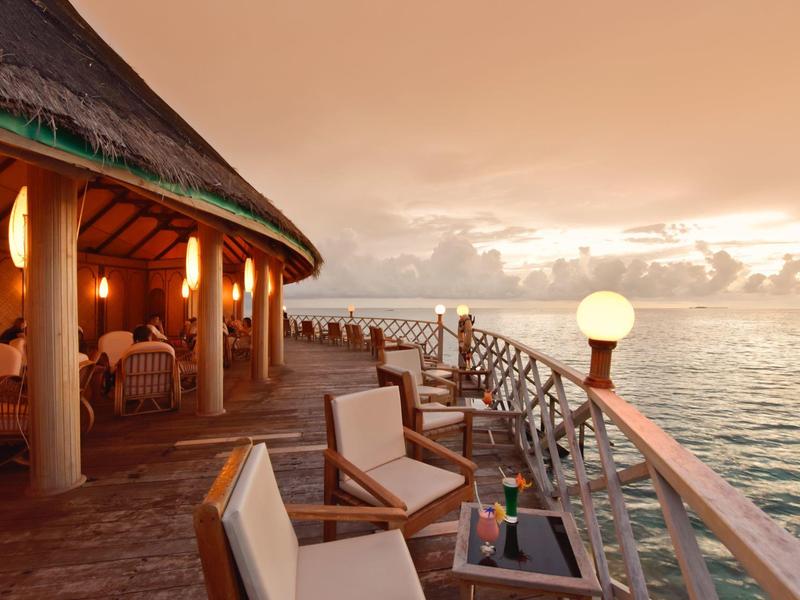 Terrassenbereich mit Holzstühlen, Tisch, Laterne und Meerblick bei Sonnenuntergang.